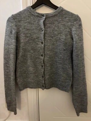 Grå kofta från H&M, XS - Mysig gråmelerad kofta från H&M i storlek XS. Koftan har rund halsringning, långa ärmar och knäppning med små knappar framtill. Ribbstickade muddar vid ärmslut och nederkant ger en klassisk look. Perfekt att slänga över en t-shirt eller topp.