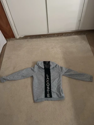 Grå hoodie från Lacoste med dragkedja - Snygg grå hoodie från Lacoste med dragkedja framtill och huva. På framsidan finns en svart panel med stor vit LACOSTE-text och krokodil-logga på huvan. Tillverkad i mjuk bomullsmix, perfekt för en chill och sportig look. Storlek S passar perfekt XS