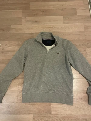 Grå halvzip sweatshirt från GANT - Snygg grå sweatshirt från GANT med halv dragkedja och liten broderad logga på bröstet. Tröjan har ribbade muddar vid ärmslut och nederkant samt hög krage. Tillverkad i mjukt bomullsmaterial som känns skönt mot huden. Helt ny inte använd en ända gång då den ej passade mig.