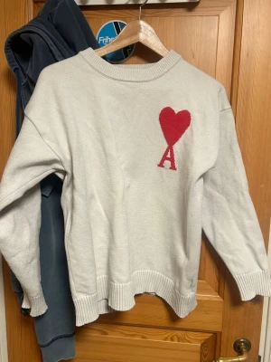 Ami Paris Sweater - Vit stickad tröja från Ami Paris med röd hjärtdetalj och bokstaven A på bröstet. Tröjan har rund halsringning, ribbade muddar och avslut. Perfekt för dig som gillar stilrena plagg med en twist.
