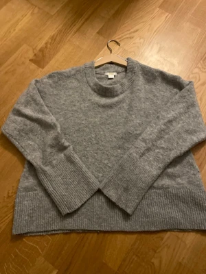 Grå stickad tröja från Gina Tricot - Mysig grå stickad tröja från Gina Tricot med rund halsringning och breda ribbade muddar vid ärmslut och nederkant. Perfekt för lager på lager och enkel att matcha till olika outfits. Materialet känns mjukt och skönt mot huden.