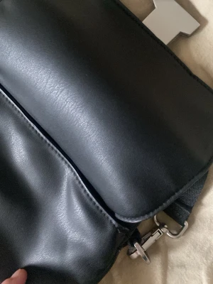 Svart axelväska med metallspänne - Snygg svart axelväska i slätt konstläder med stort T-format metallspänne framtill. Väskan har justerbar axelrem och dragkedja upptill. Insidan är fodrad i svart tyg och har en innerficka för småprylar. Perfekt för dig som gillar stilrena accessoarer.