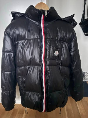 Svart dunjacka från Moncler - Snygg svart dunjacka från Moncler med glansig finish och klassisk röd, vit och blå rand längs dragkedjan. Jackan har huva, bröstficka med Moncler-logga och två sidofickor. Perfekt för kalla vinterdagar och riktigt trendig look.   QR kod samt NFC scanner funkar