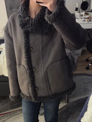 Mörkgrå teddyjacka med fårpälslook - Säljer en mörkgrå teddyjacka med fårpälslook och stora krage. Jackan har knappar framtill, två fickor och detaljer i pälsimitation. Perfekt för kalla dagar och ger en riktigt mysig vibe.