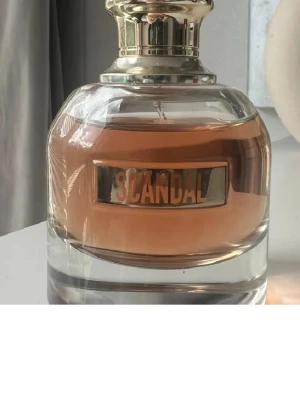 Scandal parfym från Jean Paul Gaultier - Scandal parfym med elegant rund glasflaska och guldig metallkork. Doftvätskan är ljusrosa och flaskan har en metallskylt med texten 'Scandal' på framsidan. Perfekt för dig som gillar lyxiga och trendiga dofter.