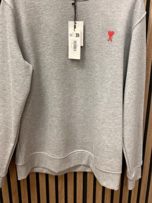 Grå sweatshirt  - Säljer en stilren grå sweatshirt med röd broderad logga i form av ett hjärta och bokstaven A på bröstet. Tröjan har rund halsringning, ribbade muddar och är tillverkad i mjuk bomullsblandning.