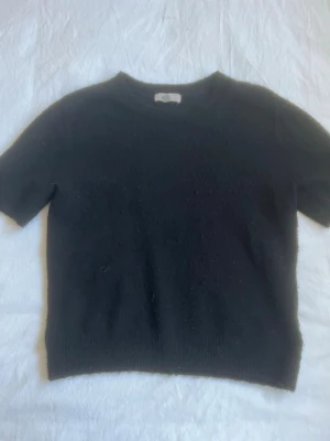 Svart stickad tröja från H&M - En enkel svart stickad tröja från H&M med korta ärmar och rund hals. Tröjan har en rak passform och ribbade kanter vid hals, ärmslut och nederkant. Perfekt att matcha med jeans eller kjol för en clean look.