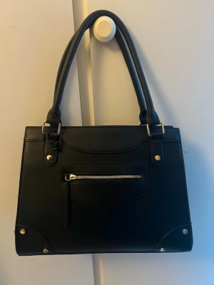 Svart handväska med gulddetaljer - Stilren svart handväska i konstläder med dubbla handtag och guldfärgade metalldetaljer. Väskan har en rak, boxig form och en praktisk ytterficka med dragkedja framtill. Perfekt för dig som gillar klassisk och enkel design. Ny pris 500