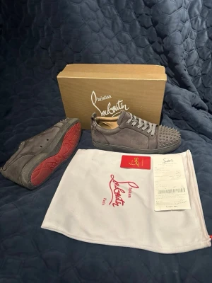 Gråa Christian Louboutin sneakers - Snygga gråa sneakers från Christian Louboutin i mocka med ikoniska röda sulor och coola nitar på tån. Skorna har lågt skaft och matchande gråa skosnören. Perfekta för dig som vill sticka ut med exklusiv design och edgy detaljer.