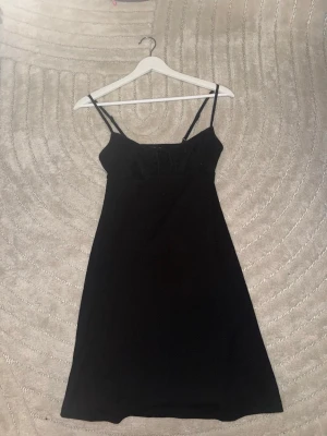 Svart kort klänning med smala band - Säljer en svart kort klänning med smala axelband och knyt framtill. Klänningen har en enkel, figurnära passform och är tillverkad i ett lätt, strukturerat tyg. Perfekt för dig som gillar minimalistisk stil och vill ha något enkelt men snyggt.