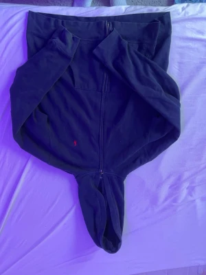 Mörkblå hoodie från Polo Ralph Lauren - Säljer en mörkblå hoodie från Polo Ralph Lauren med dragkedja framtill och klassisk huva. Tröjan har det ikoniska röda Polo-märket broderat på bröstet och är tillverkad i mjukt bomullsmaterial. Perfekt för en avslappnad och stilren look.