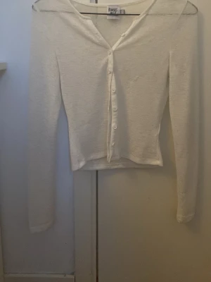 Vit cardigan från Princess Polly - Säljer en vit, långärmad cardigan från Princess Polly. Tröjan har en v-ringning och knappar hela vägen. Materialet är mjukt och tunt, vilket gör den skön att bära.