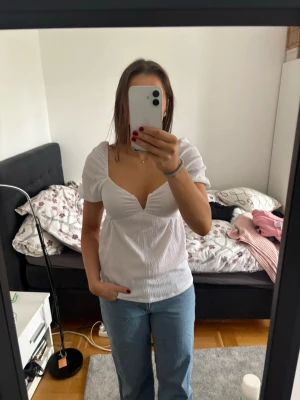 Vit v-ringad blus med puffärm - Säljer en söt vit blus med djup v-ringning och korta puffärmar. Toppen har en lätt ribbad struktur och sitter snyggt med en figurnära passform. Perfekt att styla med jeans eller kjol för en trendig look.