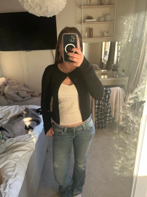 Svart cropped kofta från Gina Tricot - Stilren svart cropped kofta från Gina Tricot med rund halsringning och en knapp framtill. Koftan har långa ärmar och är tillverkad i ett mjukt, stretchigt material som passar perfekt till jeans eller kjol.