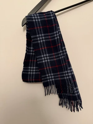 Burberry halsduk - Snygg mörkblå halsduk med klassiskt rutmönster i vitt och rött. Halsduken har fransar i ändarna och är tillverkad i cashmere och merino som känns skönt mot huden. Perfekt accessoar för att lyfta din outfit.