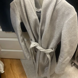 Ljusgrå kappa med huva och knytskärp från Zara - Stilren ljusgrå kappa med stor huva och knytskärp i midjan. Kappan har en mjuk, filtliknande yta och raka ärmar. Perfekt för dig som vill ha en enkel men trendig look. Passar till lager-på-lager och funkar året runt. (den är nopprig )