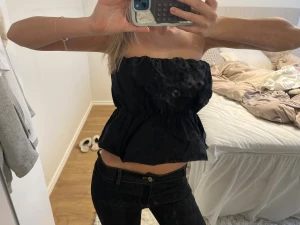 Svart bandeau tubtopp med volang - Säljer en svart bandeau tubtopp med volangkant och resår i midjan. Toppen är axelbandslös och har en luftig, croppad passform som ger en trendig look. Perfekt att matcha med jeans eller kjol för en snygg outfit.