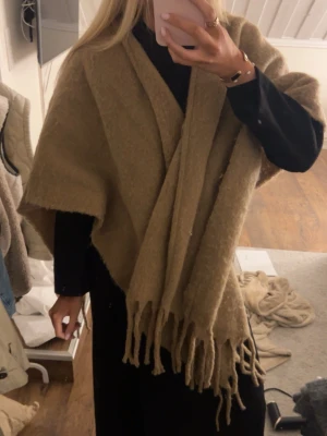 Beige stickad cape - Mysig och oversized beige cape i fluffigt material med långa fransar i kanterna. Perfekt att svepa in sig i under kyliga dagar. Enkel att styla över jacka eller tröja för en trendig lager-på-lager-look. 