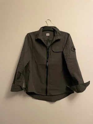 Grå overshirt från C.P. Company - Stilren grå overshirt från C.P. Company med dragkedja framtill, klassisk krage och bröstficka med lock. Ärmen har den ikoniska linsen på sidan. Tillverkad i ett lätt och slitstarkt material som passar perfekt till lager-på-lager-stil.