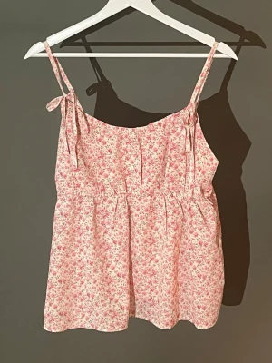 Blommig rosa linnetopp med knytband - Söt linnetopp med små rosa blommor på vit botten. Toppen har tunna axelband med knyt och en luftig, lös passform. Perfekt för varma dagar och ger en romantisk vibe till din outfit. Använd fåtal gånger, sitter bra om du har större byst!🩷