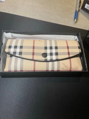 Beige plånbok med rutigt mönster - Snygg beige plånbok med klassiskt rutigt mönster i svart, vitt och rött. Plånboken har en avlång form och stängs med en svart knapp. Tillverkad i tåligt konstläder och levereras i en svart presentask. Perfekt för dig som gillar stilrena accessoarer.