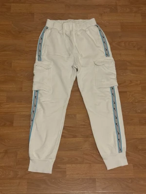 Vita cargopants med blå detaljer - Snygga vita cargopants med breda ben och coola blå ränder med svarta mönster längs sidorna. Byxorna har stora fickor på sidorna och resår i midjan med snörning för extra komfort. Perfekta för en avslappnad streetwear-look.