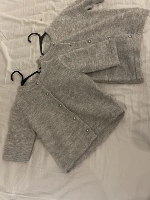 Grå stickad kofta med pärlknappar 2st - Mysig grå stickad kofta med rund hals och pärlknappar framtill. Koftan har en rak passform och långa ärmar, perfekt för lager på lager. Enkel och stilren design som passar till det mesta. På ena så är en knapp borta och den andra så är alla på det är 146 och 140 i stolek 