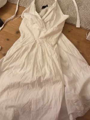 Vit omlottklänning med v-ringning - Snygg vit klänning med omlottliv och v-ringning. Klänningen har en utställd kjol och är ärmlös, perfekt för varma dagar. Tillverkad i ett lätt bomullstyg som ger en luftig känsla.