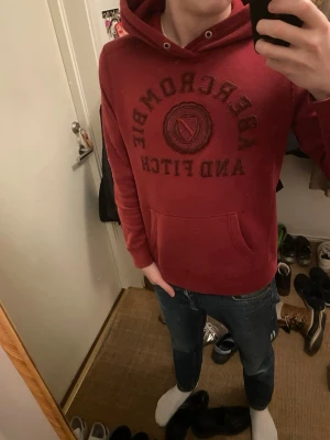 Röd hoodie från Abercrombie & Fitch - Snygg röd hoodie från Abercrombie & Fitch med stor logga och text på bröstet. Klassisk känguruficka framtill och justerbar huva med dragsko. Tillverkad i mjukt material som känns skönt mot huden. Perfekt för en avslappnad och trendig look.