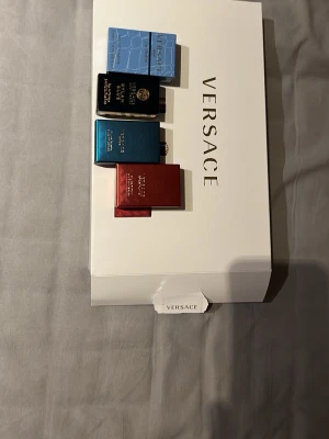 Versace parfymset med fyra miniflaskor - Snyggt parfymset från Versace med fyra miniflaskor i olika färger: ljusblå, svart, mörkblå och röd. Flaskorna har elegant design med guld- och blå detaljer och levereras i en vit Versace-box. Perfekt för dig som vill testa flera dofter från ett lyxigt märke. Väldigt fina parfymer aldrig andvända köpte dem i början av februari och har inget behov av dem längre!