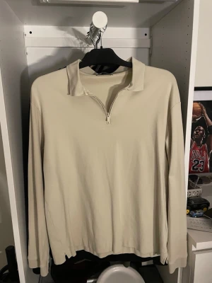 Beige långärmad tröja med dragkedja - Stilren beige långärmad tröja med halv dragkedja och klassisk krage. Perfekt för en avslappnad men ändå snygg look. Perfekt om man vill klä upp sig utan att stå ut för mycket. Storlek M. Aldrig använd