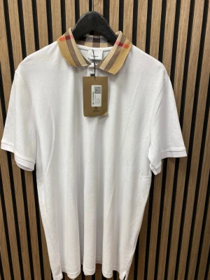 Vit t-shirt med rutig krage  - Säljer en stilren vit t-shirt med klassisk rutig krage i beige, svart och rött. T-shirten har korta ärmar och är gjord i mjuk bomull. Perfekt för dig som vill ha en clean look med en twist.