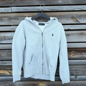 Grå hoodie från Polo Ralph Lauren - Ljusgrå hoodie från Polo Ralph Lauren med dragkedja framtill och klassisk huva. storlek M men sitter som en S nästan förliten för mig som är 178. finns även två fläckar (se bild) en på insidan så den syns inte när den sitter på och en på armen som borde gå o få bort. 