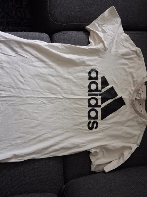 Vit Adidas t-shirt med logga - Säljer en vit t-shirt från Adidas med stor svart logga på bröstet. Klassisk rund hals och korta ärmar, perfekt för dig som gillar sportiga och stilrena plagg. Materialet är mjuk bomull som känns skönt mot huden.