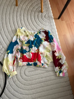 Färgglad blommig blus med lång ärm - Blus med färgstarkt blommönster i blått, rött, gult och rosa. Modellen har långa puffärmar med resår vid ärmslut och midja. Materialet ser ut att vara lätt och luftigt, perfekt för en trendig look. Rund halsringning och avslappnad passform.
