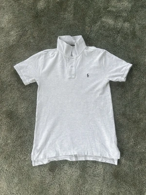 Ljusgrå piké från Ralph Lauren - Snygg ljusgrå pikétröja från Ralph Lauren med klassisk krage och två knappar framtill. Tröjan har korta ärmar och den ikoniska lilla loggan broderad på bröstet. Tillverkad i mjuk bomull som känns skön mot huden.