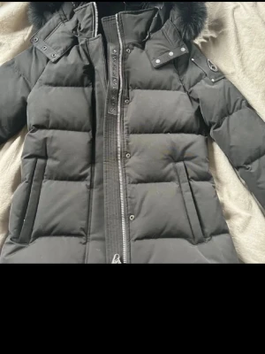Svart dunjacka från Moose Knuckles - Säljer en svart dunjacka från Moose Knuckles med pälskantad huva och logga på ärmen. Jackan har quiltad design, hög krage och snygga detaljer med tryckknappar på axlarna. Perfekt för kalla vinterdagar och riktigt varm.