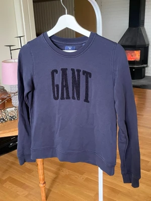 Mörkblå sweatshirt från GANT - Mörkblå sweatshirt från GANT med stor, svart broderad logga på bröstet. Tröjan har rund halsringning, långa ärmar och ribbade muddar vid ärmslut och nederkant. Tillverkad i mjukt bomullsmaterial som är skönt att ha på sig.