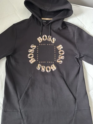 Svart hoodie från Hugo Boss - Svart hoodie från Hugo Boss med stor logga i beige och vitt på bröstet där det står 'BOSS' i ett cirkelformat mönster. Klassisk huva med dragsko och känguruficka framtill. Tillverkad i mjukt bomullsmaterial, perfekt för en avslappnad och trendig look.