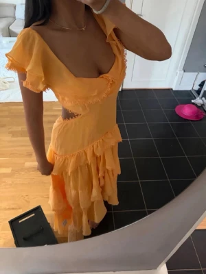  volangklänning med cut out - Säljer en slutsåld lång gul klänning med volanger och cut out-detaljer i midjan. Klänningen har korta volangärmar, omlott och knyt i ryggen samt v-ringad hals. Perfekt för sommarens alla tillfällen. Materialet känns lätt och luftigt. Size S-M går att knyta och tajta till där bak. Ljus orangel/coral färg