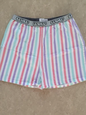 Shorts - Säljer ett par färgglada randiga shorts från Wow med elastisk midja och logotyp på resåren. Shortsen har vertikala ränder i lila, mintgrön, rosa och vit. Perfekta för varma dagar och chill stunder.