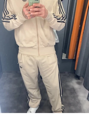 Beige Adidas loungewear dress helt nu Smal  - Säljer ett beige loungewear set från Adidas med klassiska svarta ränder på ärmar och ben. Setet består av en ziptröja med hög krage och matchande byxor med dragsko i midjan. Båda delarna har Adidas logga framtill. Perfekt för chill dagar hemma. Helt ny nypris stadium 849 och mjukis 700 säljes för tusen 