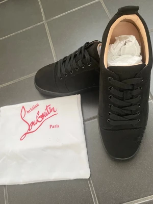 Svarta sneakers från Christian Louboutin - Snygga svarta sneakers från Christian Louboutin med klassisk röd sula och diskret logga. Skorna har en låg profil, svarta skosnören och ovandel i mocka. Insidan är beige och de har en exklusiv vibe som sticker ut.