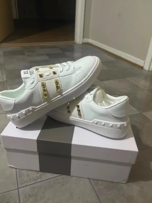 Vita Valentino sneakers med nitar - Snygga vita sneakers från Valentino med guldiga pyramidformade nitar på sidorna och detaljer i vitt läder. Skorna har lågt skaft och coola textilpaneler med nitar som ger en edgy vibe. Perfekta för dig som vill sticka ut med din stil.