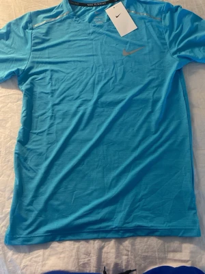 Blå Nike löpartröja herr - Nike löpartröja i klarblått funktionsmaterial med korta ärmar och rund hals. Tröjan har en diskret Nike-logga på bröstet och reflekterande detaljer på axlar och rygg. Perfekt för löpning eller träning, lätt och ventilerande.