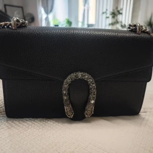 Svart handväska i läder - Säljer en svart  handväska i läder med silverfärgad kedja och ikoniskt spänne med ormdetaljer. Väskan har ett ljust innerfoder och praktiskt innerfack med dragkedja. Perfekt för dig som vill ha en lyxig och stilren accessoar.
