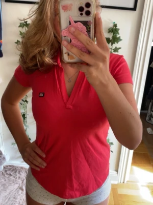 Rosa kortärmad polotopp från Flash Leisure - Snygg rosa polotopp från Flash Leisure med korta ärmar och liten logga på bröstet. Toppen har en klassisk krage och v-ringad öppning framtill. Perfekt för en sportig och avslappnad look.