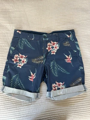 blå shorts  - Snygga blå shorts med tropiskt blommönster i rosa och vitt samt gröna blad. Shortsen har uppvikta benslut, sidofickor och bakfickor. Perfekta för sommarens alla äventyr och har en avslappnad passform.