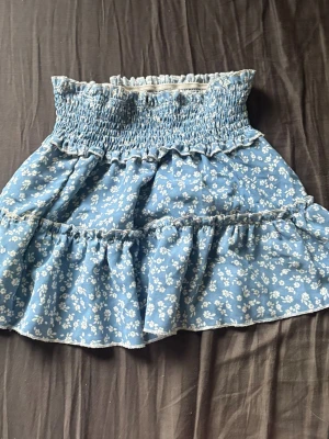 Blå blommig volangkjol - Söt blå kortkjol med vita små blommor och volangdetaljer. Kjolen har en smockad midja som ger en bekväm och stretchig passform. Perfekt för varma dagar och enkel att matcha med olika toppar. Hittar ingen storlekslapp men vill minnas att den är stl xs