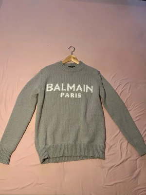 Grå stickad tröja från Balmain - Snygg grå stickad tröja från Balmain Paris med vit logga framtill. Tröjan har rund halsringning, långa ärmar och ribbstickade muddar vid ärmslut och nederkant. Perfekt för dig som gillar exklusiva och trendiga plagg.